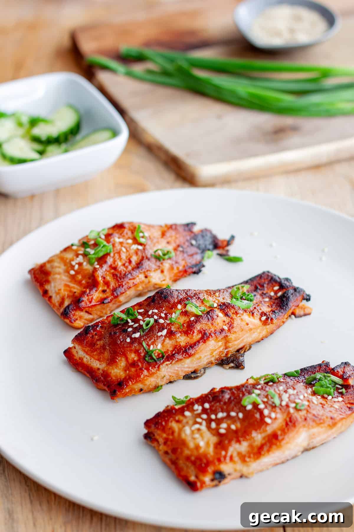 A platter of miso salmon on a butcherblock countertop with scallions and cucumbers in the background.