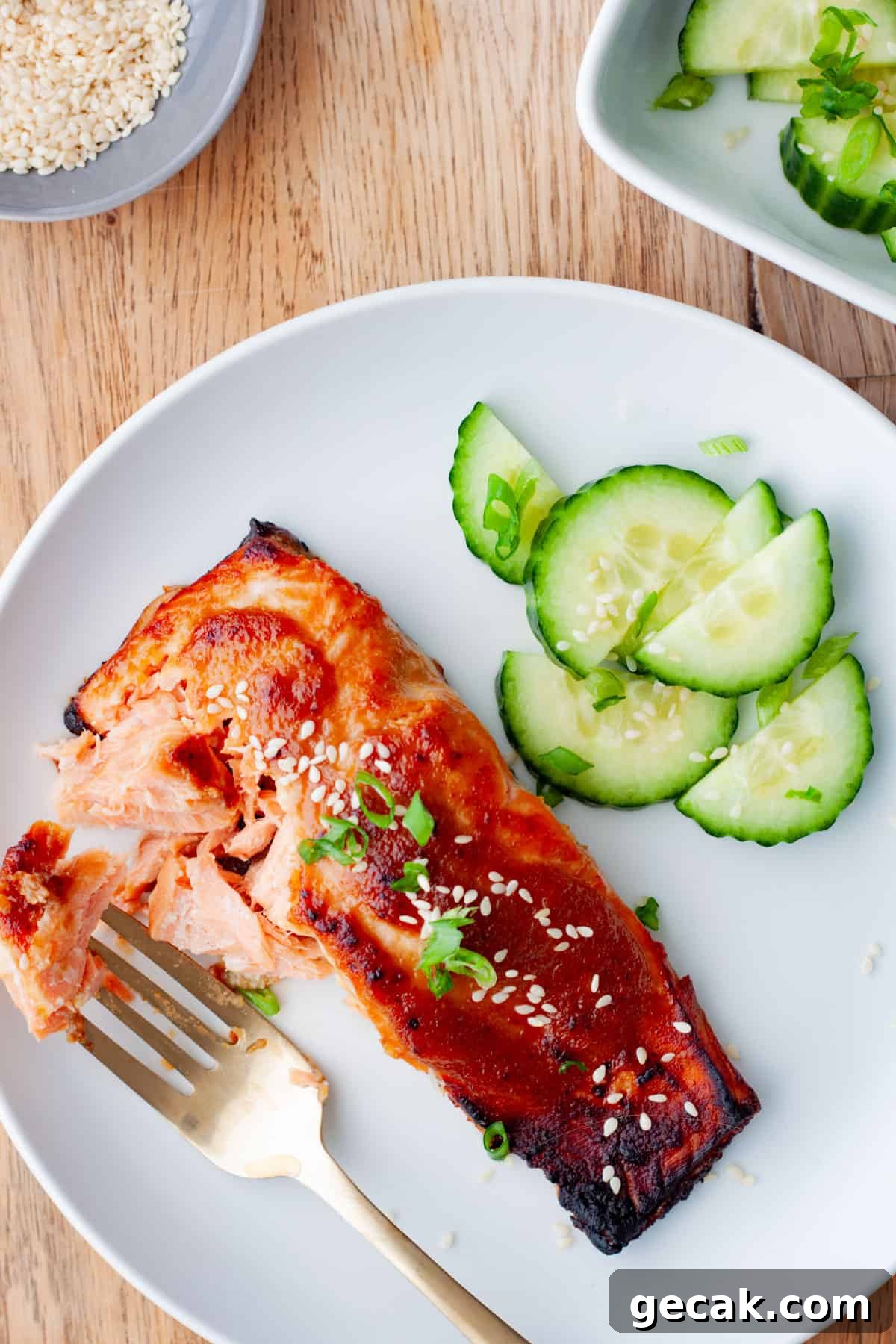 A fork taking a bite out of miso salmon with sliced cucumbers to the side.