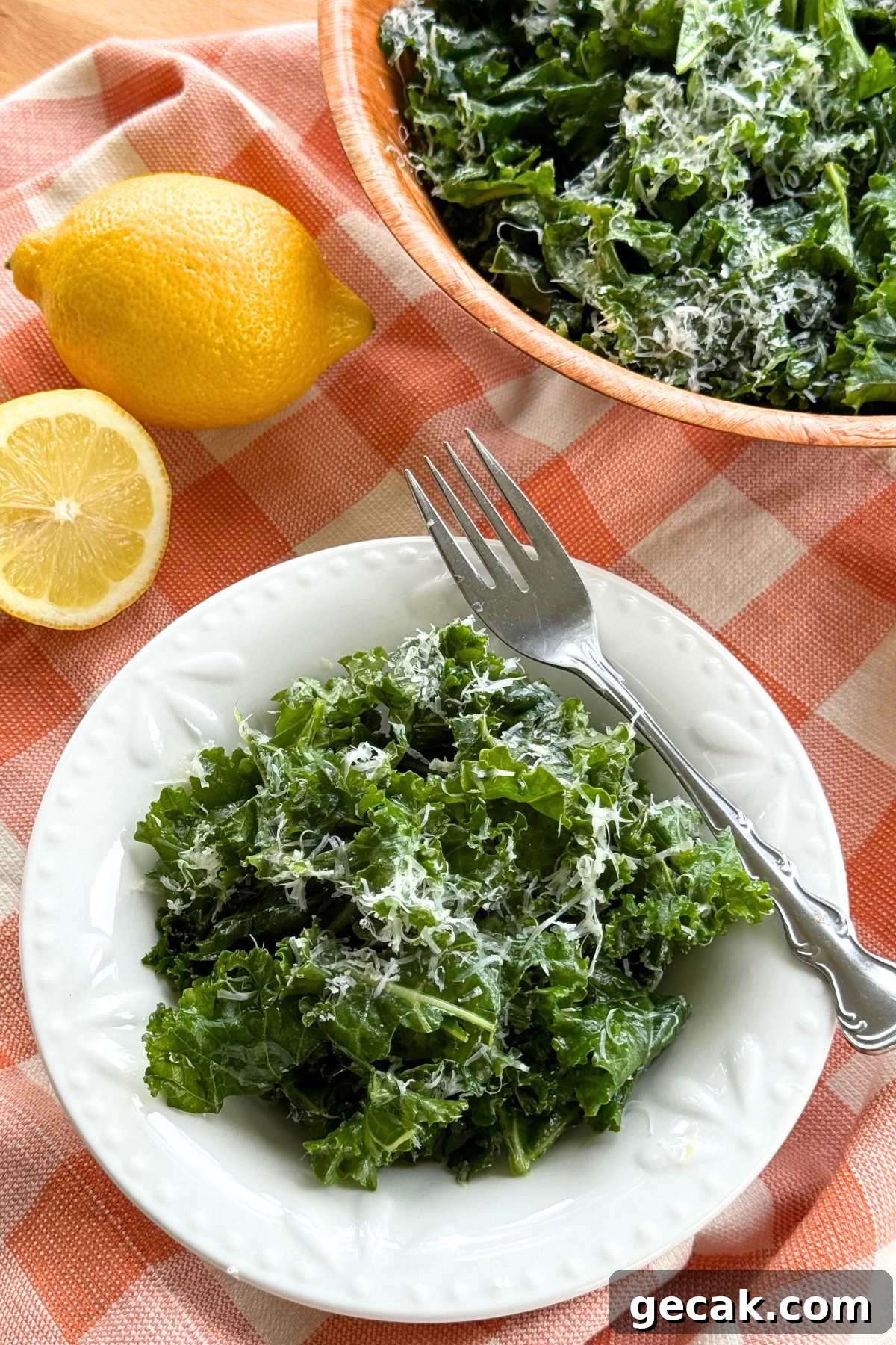 Massaged kale salad topped with finely grated Parmesan cheese and lemon dressing in a white bowl on a orange checkered background with a lemon and serving bowl with more behind it.