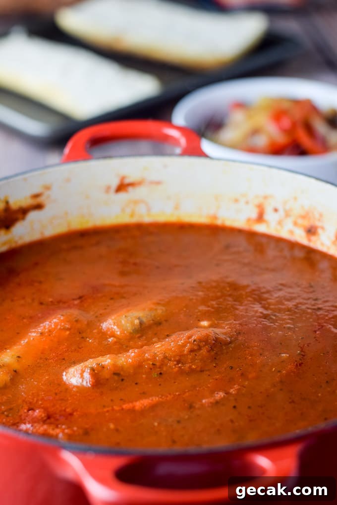 Marinara cooked with sausages in it with a bowl of cooked onions and peppers and bread on a pan in the background