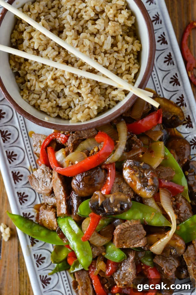 A serving platter of flank steak stir fry next to a bowl of brown rice and wooden chop sticks.