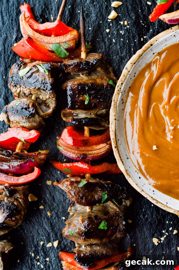 Flank steak, red onions and red bell peppers on wooden skewers with a bowl of peanut sauce next to them.