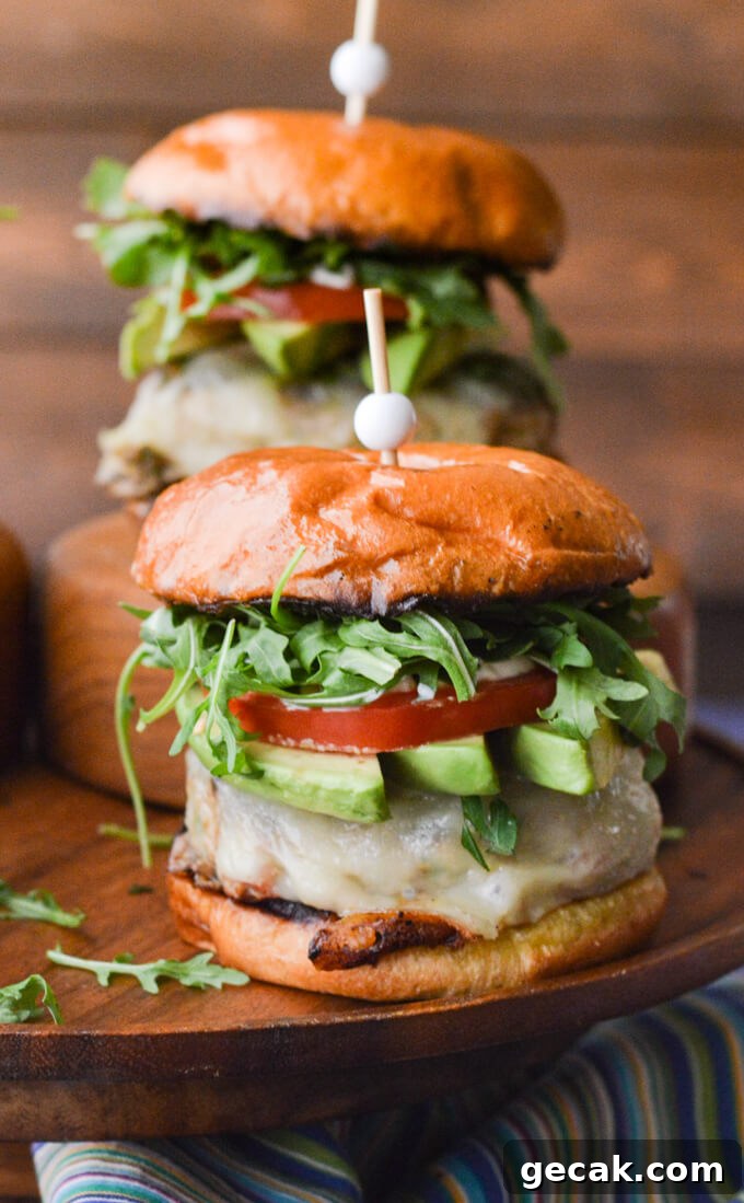 A front view of two turkey burgers with avocado slices, tomato and arugula sitting on a wooden tray with a blue striped napkin in front.
