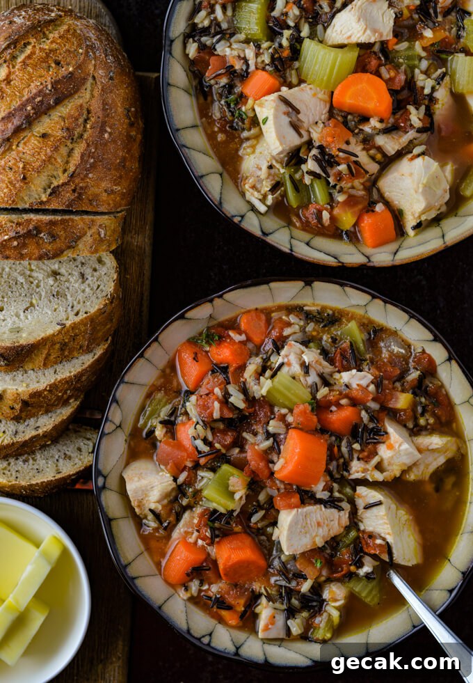 Two bowls of wild rice chicken soup loaded with carrots, onions and celery. A sliced load of bread with a small white bowl of butter cubes sit next tot he soup.