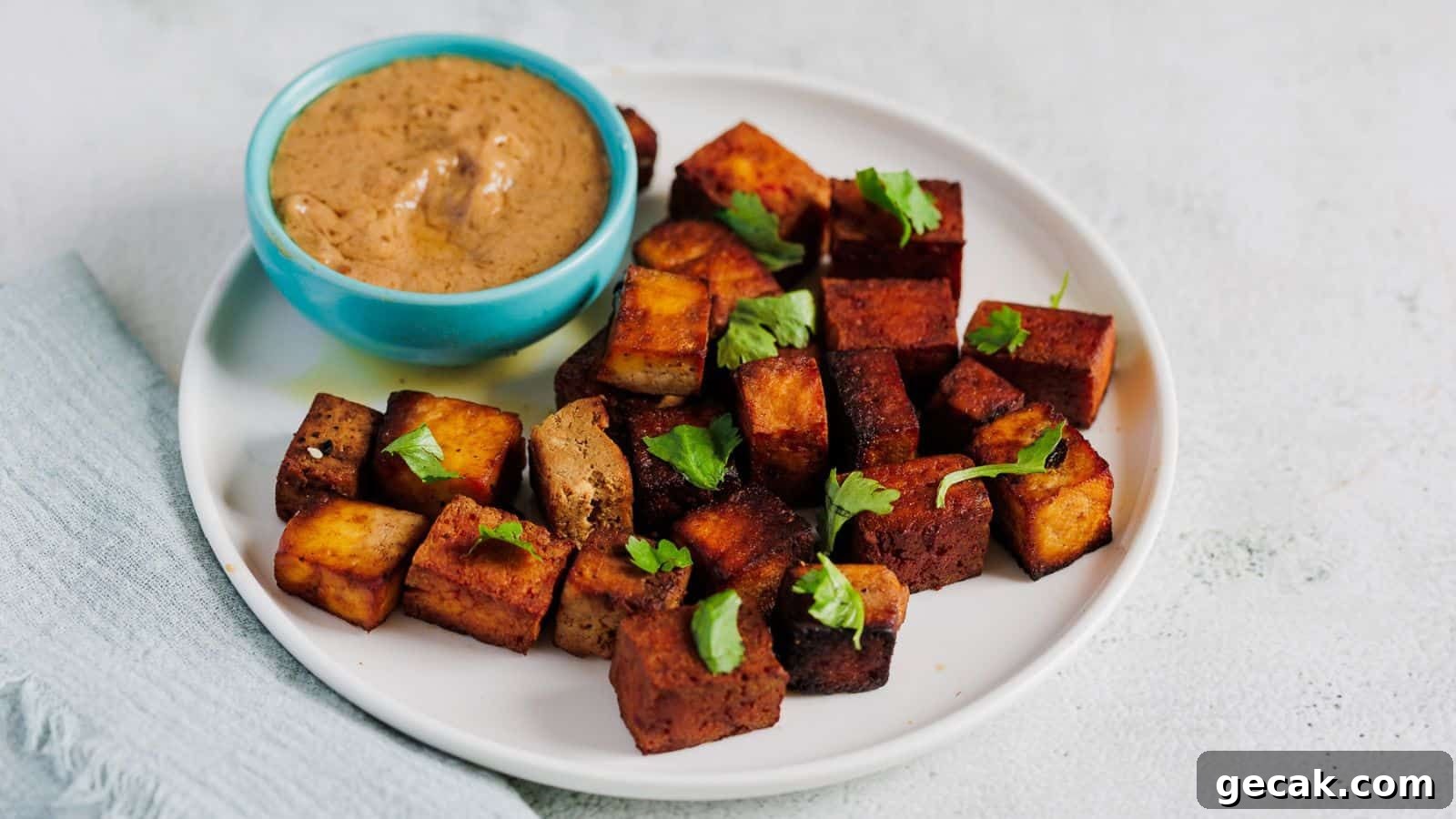 A white plate with cubes of cooked tofu garnished with herbs, served with a small bowl of creamy brown dipping sauce.