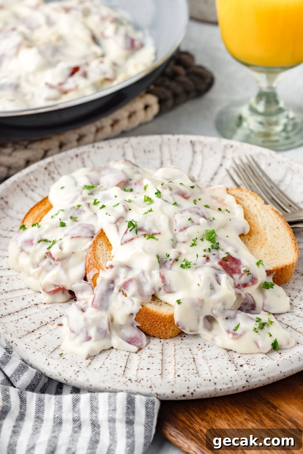 Two slices of white bread topped with creamy chipped beef gravy, garnished with chopped parsley, are served on a speckled plate with a fork and a glass of orange juice nearby.