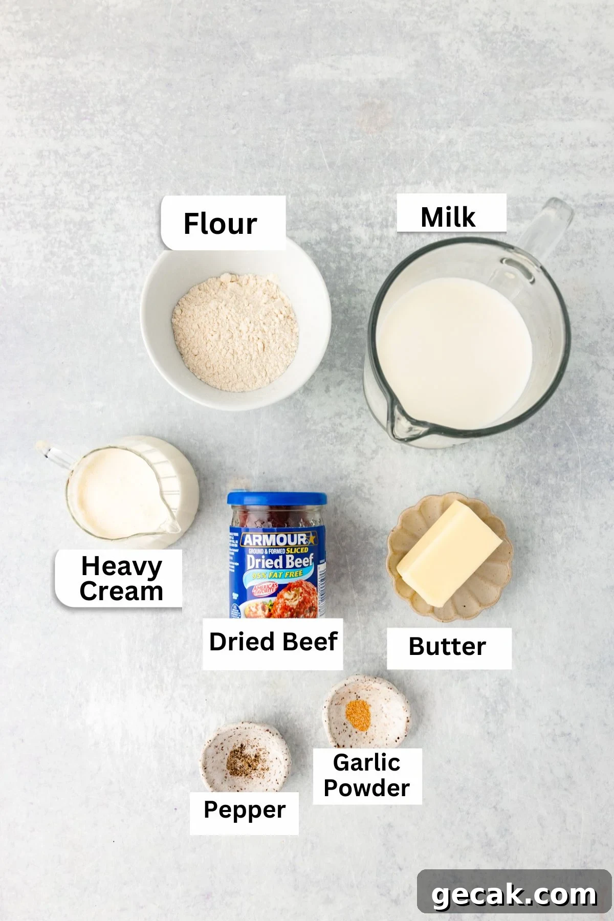 Ingredients for dried beef gravy are arranged on a light surface: flour, milk, heavy cream, a jar of dried beef, a stick of butter, and small bowls of pepper and garlic powder, each labeled.
