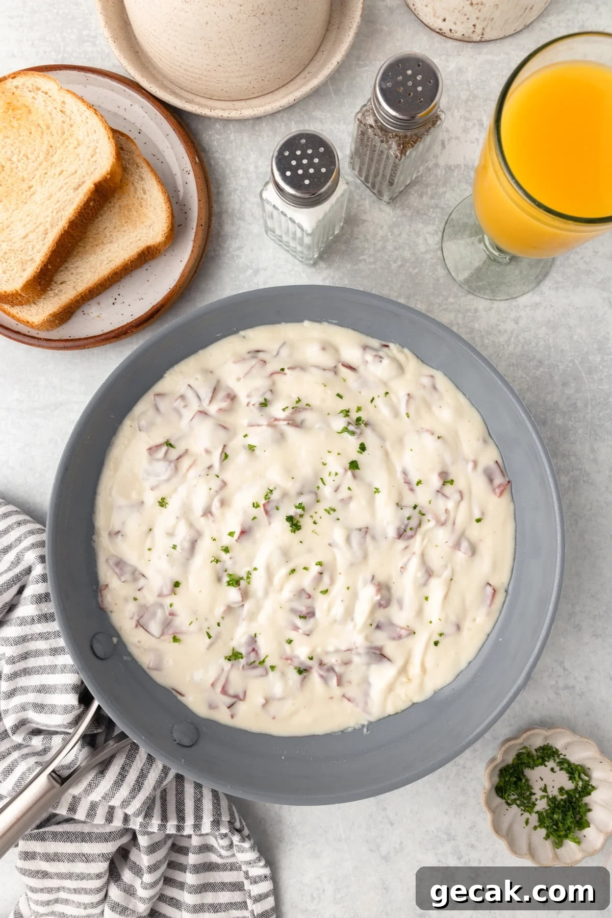 A skillet of creamy sausage gravy garnished with chopped herbs sits on a light countertop, surrounded by toast, a glass of orange juice, salt and pepper shakers, and a small bowl of minced parsley.
