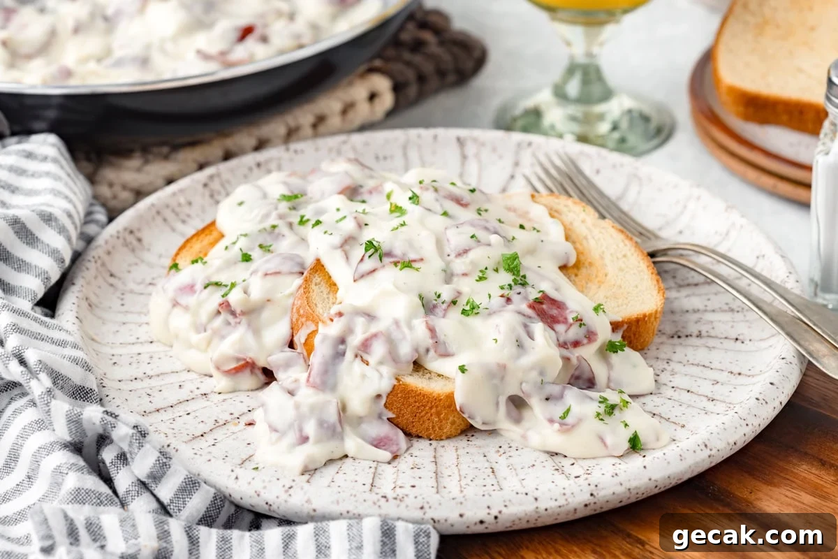 Two slices of white bread topped with creamy chipped beef in white sauce, garnished with chopped parsley, served on a speckled plate with a fork beside it. A striped napkin and glass are nearby.