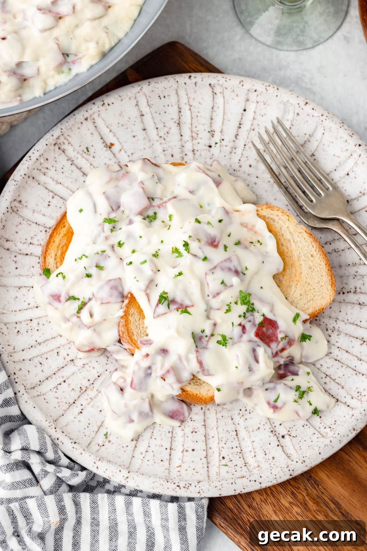 Two slices of toasted bread topped with creamy chipped beef and garnished with chopped parsley are served on a speckled plate, with a fork on the side and a striped napkin underneath.