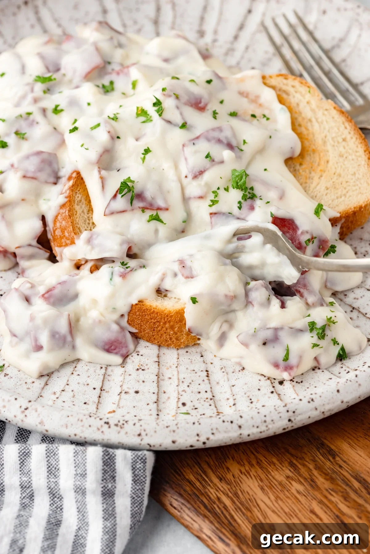 Slices of bread topped with creamy chipped beef and white sauce, garnished with chopped parsley, served on a speckled plate with a fork nearby.