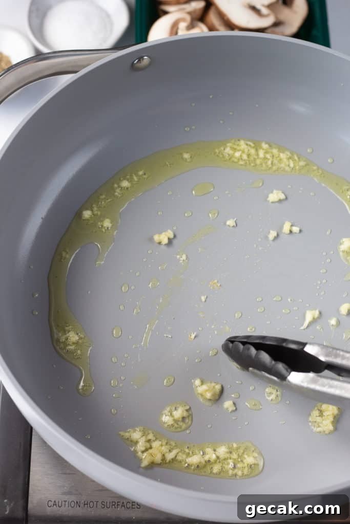 20-Minute Sautéed Zucchini and Mushrooms Recipe 4 oil and minced garlic in a gray pan being spread by tongs