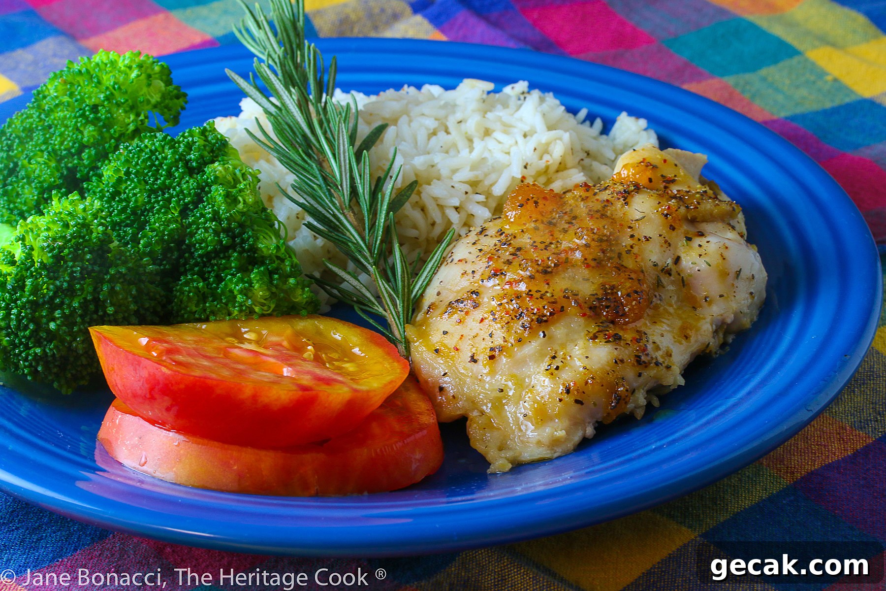 Golden chicken with an apricot-wine glaze served with sliced red tomatoes, rice, and broccoli on a blue plate placed on a colorful plaid napkin © 2023 Jane Bonacci, The Heritage Cook.