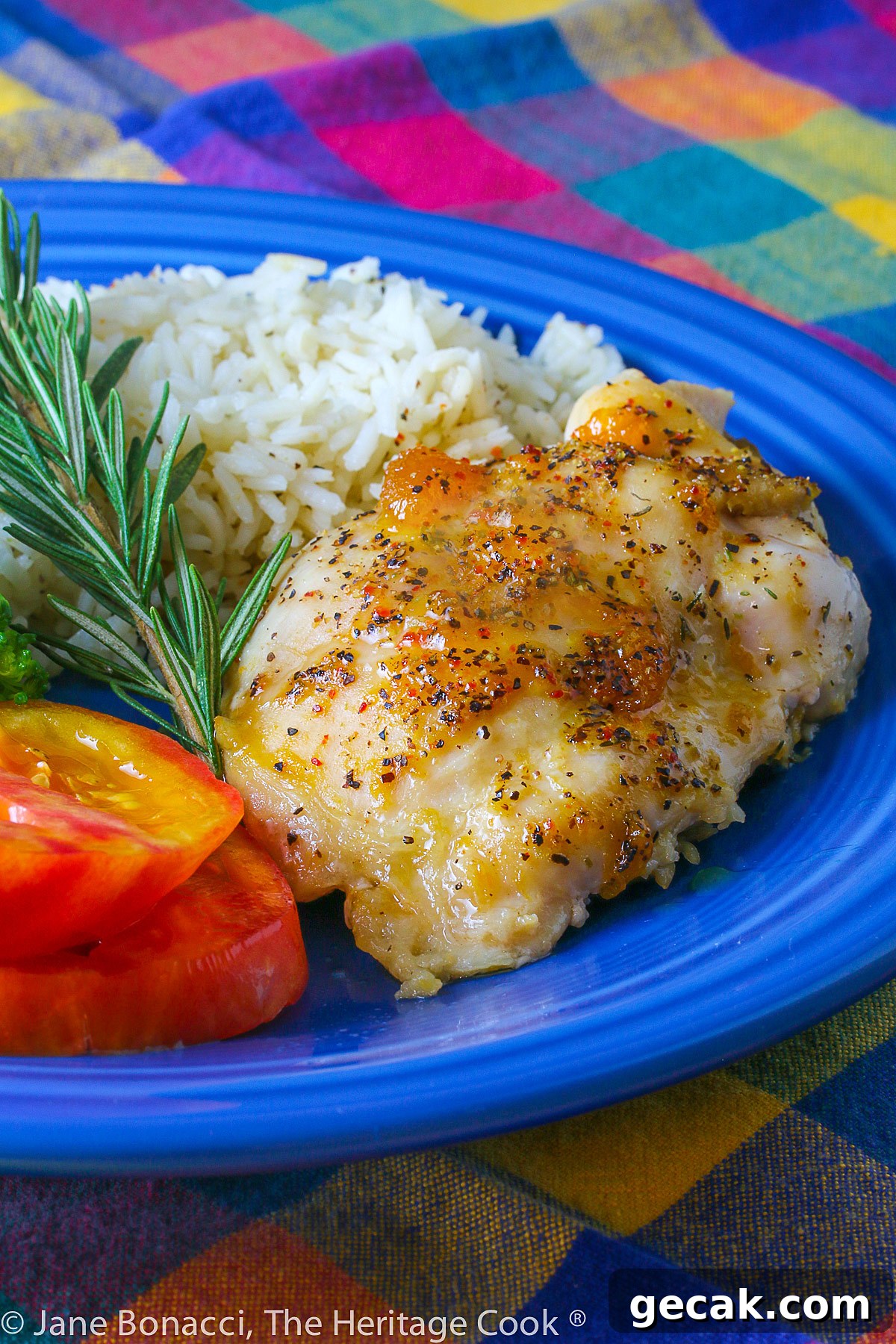 Golden chicken with an apricot-wine glaze served with sliced red tomatoes, rice, and broccoli on a blue plate placed on a colorful plaid napkin © 2023 Jane Bonacci, The Heritage Cook.