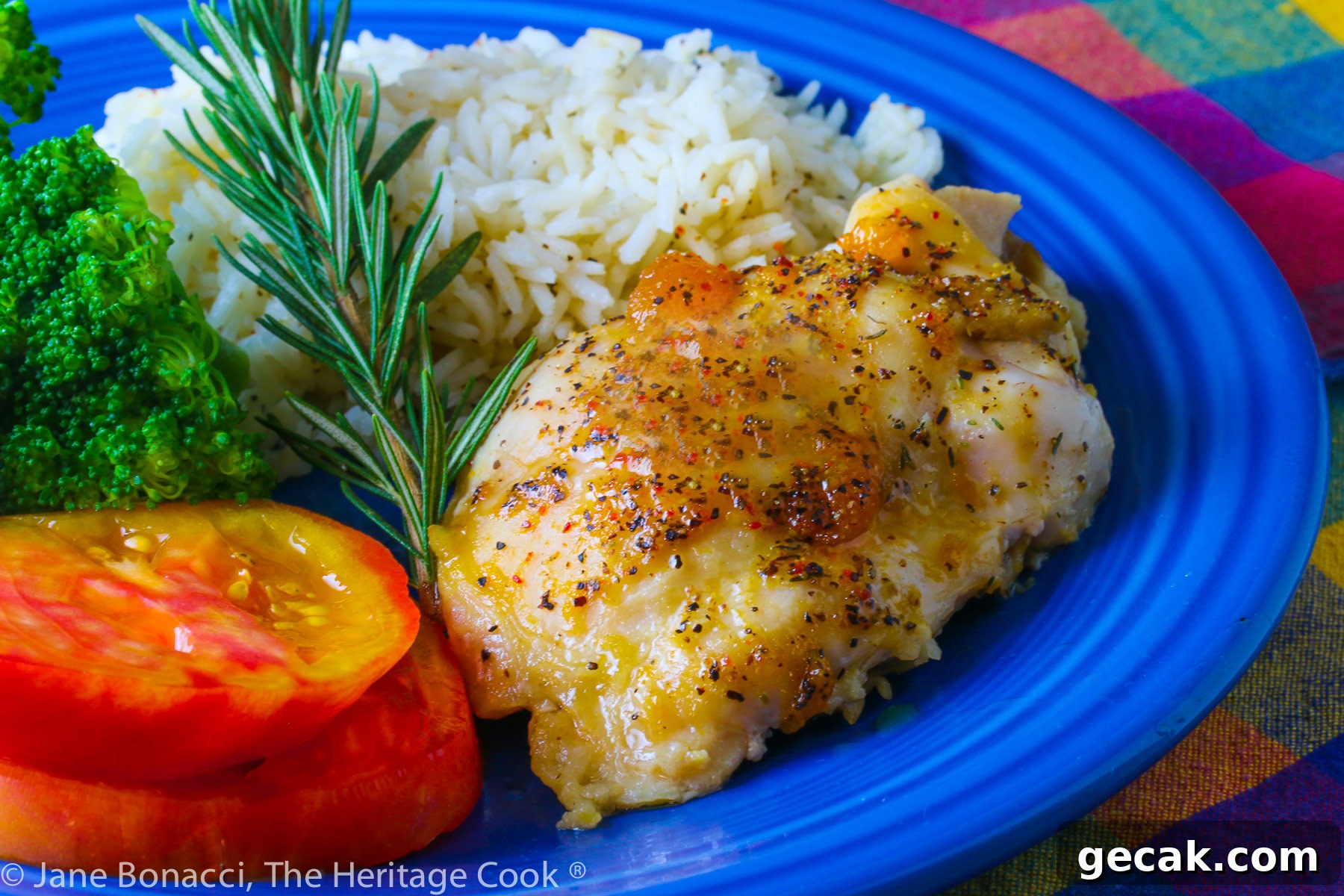 Golden chicken with an apricot-wine glaze served with sliced red tomatoes, rice, and broccoli on a blue plate placed on a colorful plaid napkin © 2023 Jane Bonacci, The Heritage Cook.