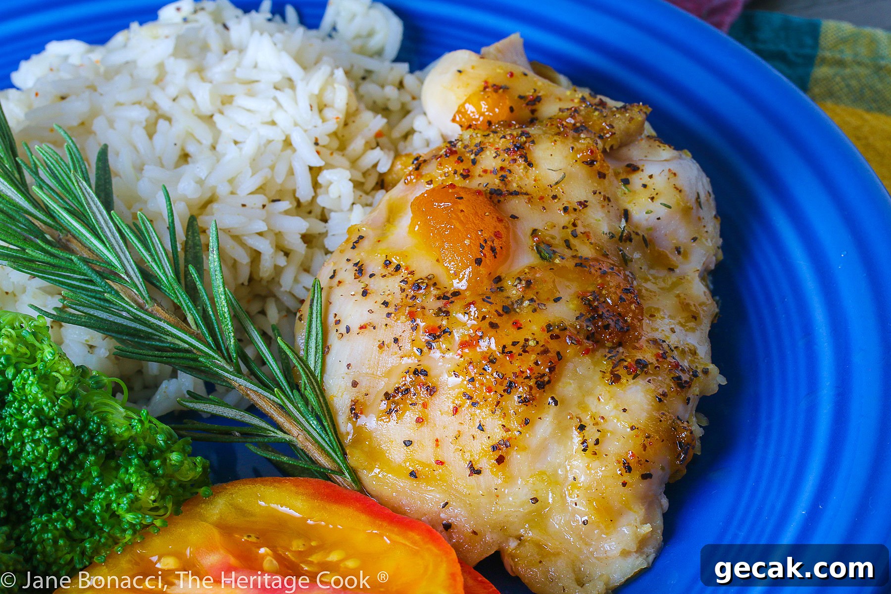 Golden chicken with an apricot-wine glaze served with sliced red tomatoes, rice, and broccoli on a blue plate placed on a colorful plaid napkin © 2023 Jane Bonacci, The Heritage Cook.
