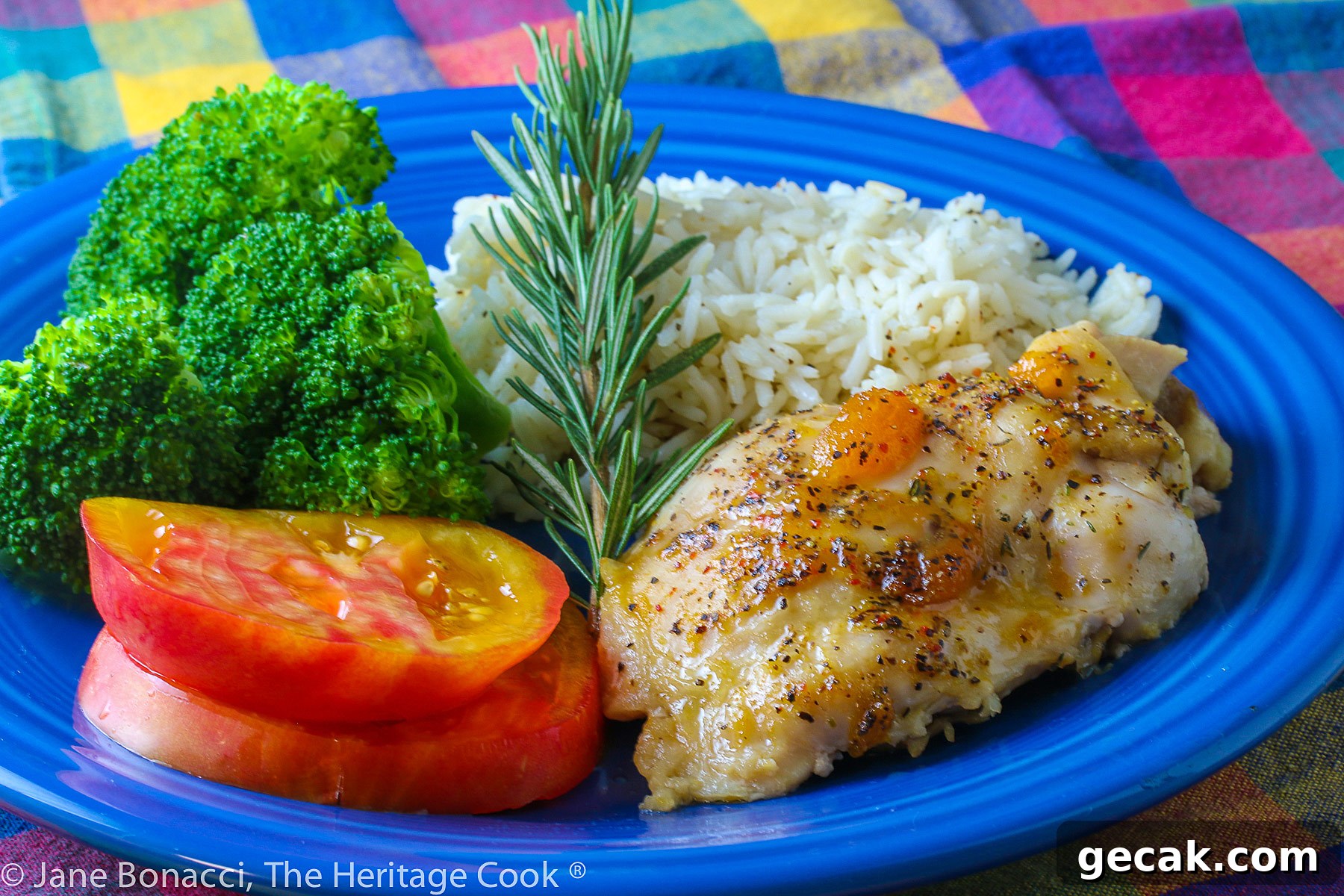 Golden chicken with an apricot-wine glaze served with sliced red tomatoes, rice, and broccoli on a blue plate placed on a colorful plaid napkin © 2023 Jane Bonacci, The Heritage Cook.