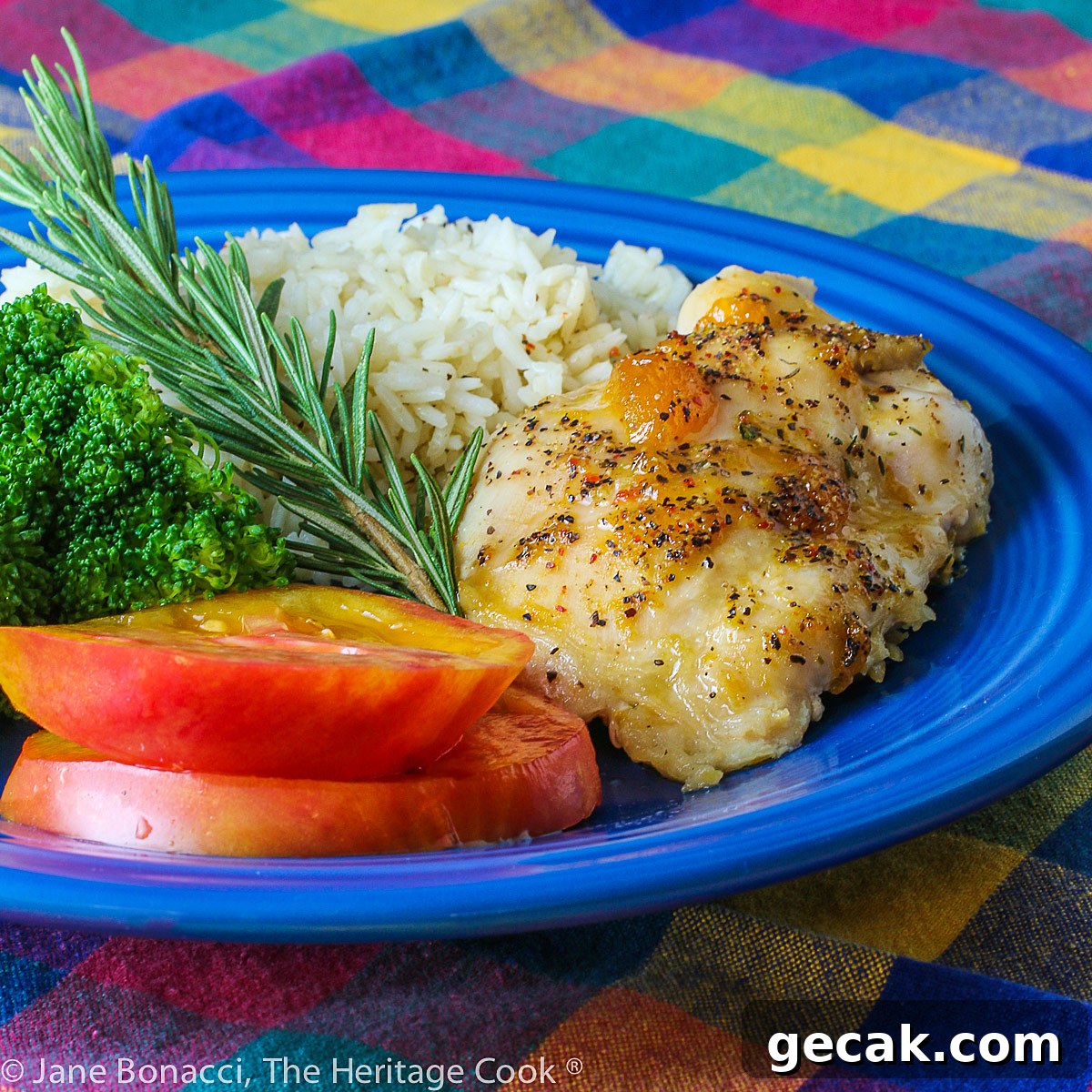 Golden chicken with an apricot-wine glaze served with sliced red tomatoes, rice, and broccoli on a blue plate placed on a colorful plaid napkin © 2023 Jane Bonacci, The Heritage Cook.